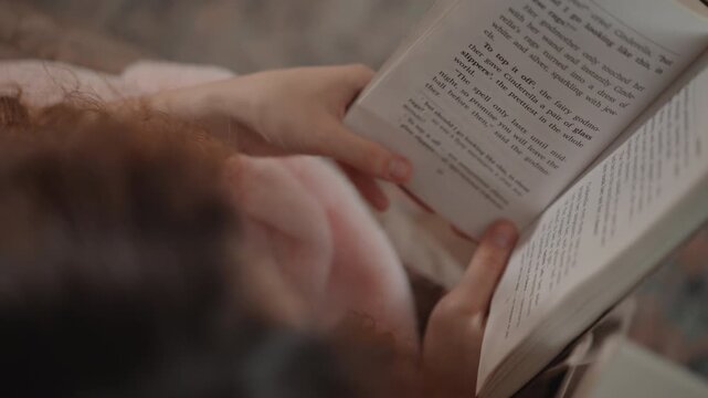 Overhead intimate view of adult hands thumb-through and turn pages of small paperback book, visible page curl and finger motion indicating active reading and page flipping; soft warm indoor lighting