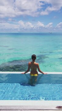 Travel vacation luxury resort idyllic overwater bungalow villa woman relaxing by infinity pool. Elegant beautiful lady in yellow bikini. Raw unedited vertical video of real woman model without makeup.