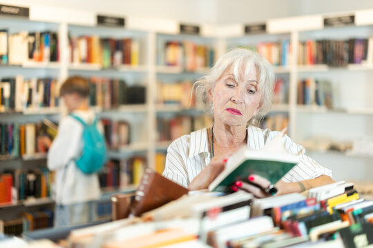 Attentive elderly woman browsing through literature on display counter in cozy bookshop