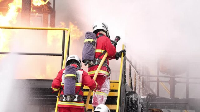 Firefighter fire hose smoke flame industrial rescue emergency training responder, crew climbing metal stairway spraying water on refinery facility blaze, thick steam, heat, urgent teamwork response