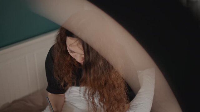 Young woman seated on bed leans forward with bowed head and long curly hair falling over face while holding pillow in her lap, creating quiet inward mood in soft bedroom light, with woman, bed