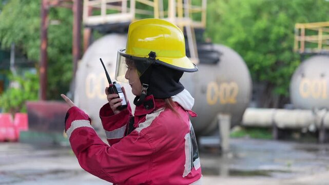 Firefighter female helmet protective suit two way radio industrial tank refinery safety worker communication during emergency drill and hazardous response at fuel storage plant, wet pavement focus