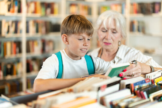 Smiling grandmother with grandson holding a book near display stand in public library