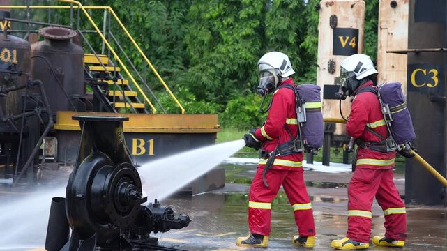 Firefighter fire hose water spray training industrial safety drill, responder team in protective suit and breathing apparatus cooling burning equipment during emergency operation at plant yard