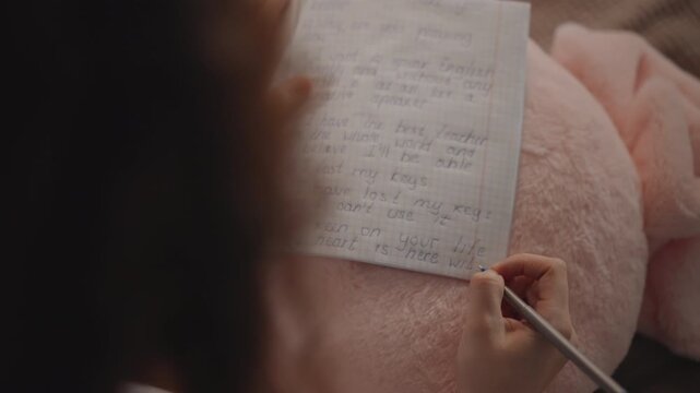 Overhead close view shows person writing fresh notes on lined paper resting on soft pink cushion in lap, right hand guiding pen while left hand steadies page in calm focused indoor setting