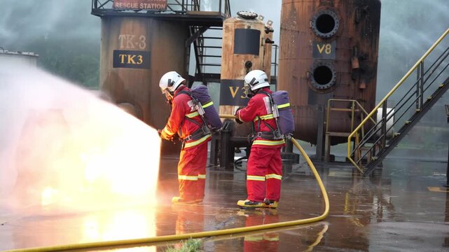 Firefighter fire hose water spray foam industrial training emergency drill scene with two responders in protective gear suppressing fuel blaze on wet concrete near refinery tank smoke