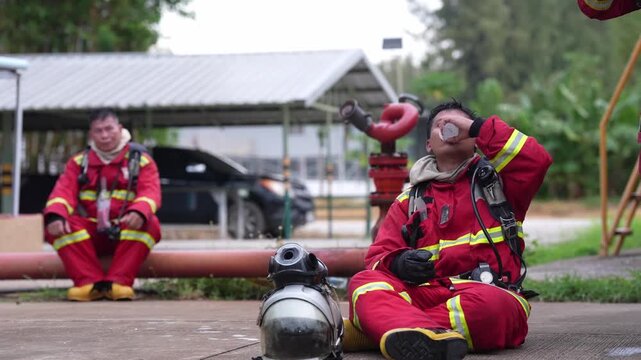 Firefighter firefighting in protective suit with breathing apparatus during training rest by hydrant after rescue drill, exhaustion recovery on pavement, drinking water beside air cylinder and hose