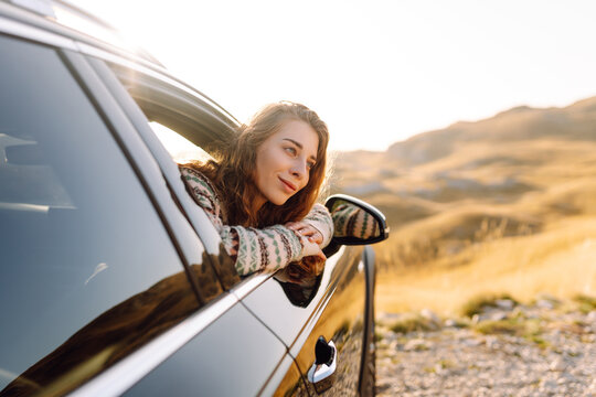 Female traveler leans out car window and enjoys mountain views. Woman on road trip experiences freedom at sunset. Concept: transportation, leisure. Active lifestyle.
