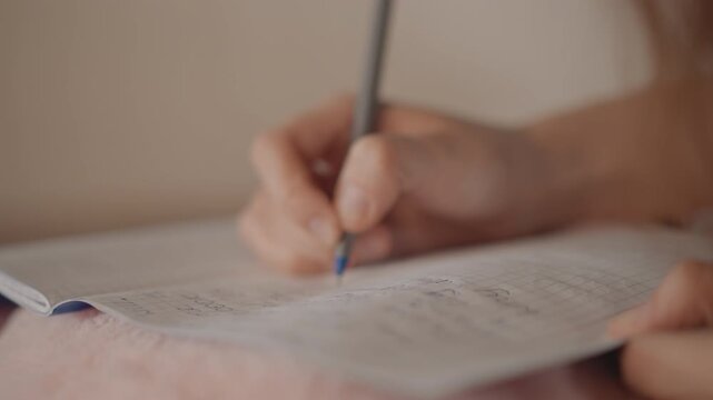 Right hand holding pencil makes downward strokes on lined notebook while left hand steadies page; visible motion blur on fingers and pencil tip shows active penmanship, pressure on paper, and
