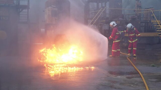 Firefighter battling industrial refinery fire with hose and foam during emergency safety drill, protective suit crew controlling intense fuel blaze, smoke and flame on wet factory ground