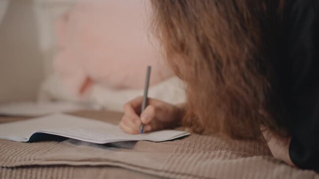 Close view of young person lying on bed and writing on paper with pen, hand making deliberate strokes while relaxed bedding, soft daylight and quiet bedroom atmosphere support focused personal routine