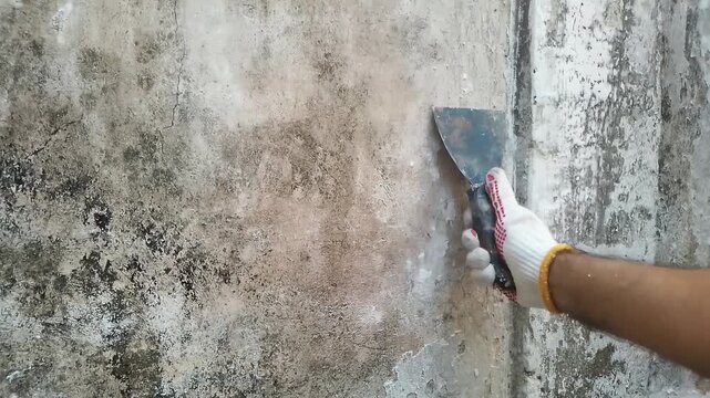 Worker using a putty knife to scrape peeling paint and plaster from a damaged wall during home renovation.

