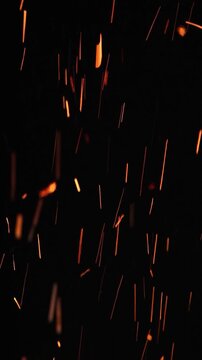 Glowing sparks rising from burning firewood at night, illuminating the darkness with warm, flickering light. Close-up and slow-motion shot on black background