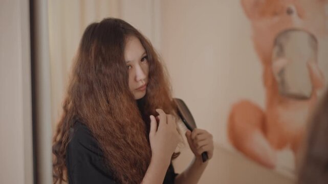 Adult woman gently detangling long curly hair before mirror, using paddle brush and hand to guide strands, calm focused grooming scene in warm dressing room with soft light, with woman, hair, brush