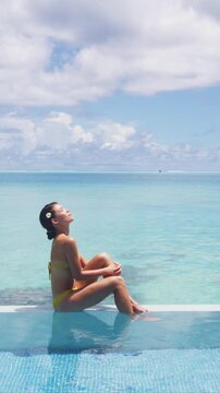Summer sunbathing bikini woman in infinity pool of luxury resort hotel relaxing in idyllic overwater ocean suite. Suntan skin care. Raw vertical video of real unretouched woman model without makeup