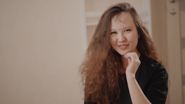 Young woman adjusts long voluminous curly hair with both hands while expressions shift from smile to playful squint and slight pout, creating lively beauty portrait with warm indoor light