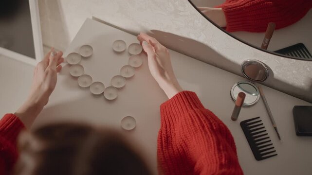 High-angle view shows hands in red sweater placing clear glass marbles into curved circle on beige desk, adjusting positions with gentle motion blur, soft light, calm indoor composition