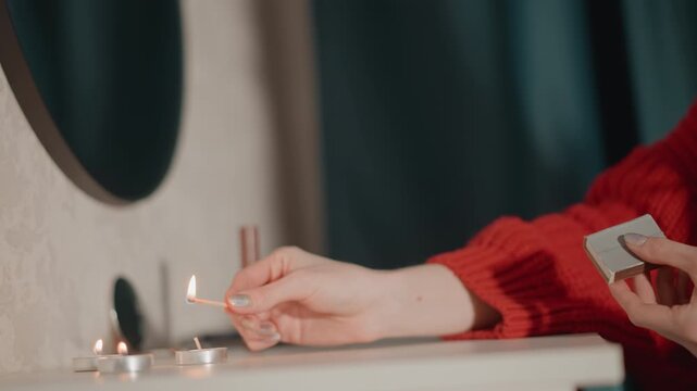 Close view shows hand in red knit sweater holding lit match above small pile of pills and blister pack on bathroom countertop, with flame glowing beside mirror and curtain in indoor setting