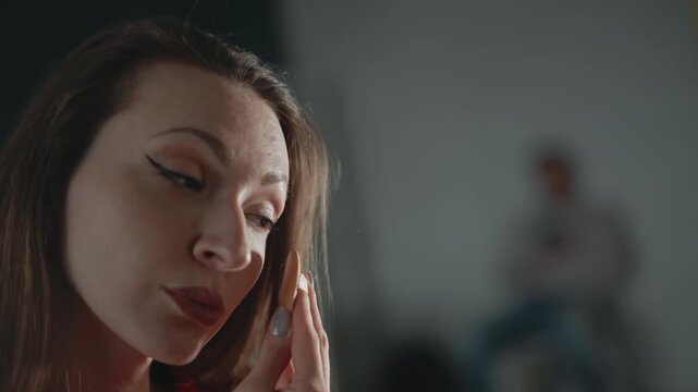 Young woman in red knit sweater gently wipes lower eyelid and cheek with cotton pad, making soft upward motions while removing makeup residue, eyes closed, expression calm, indoors