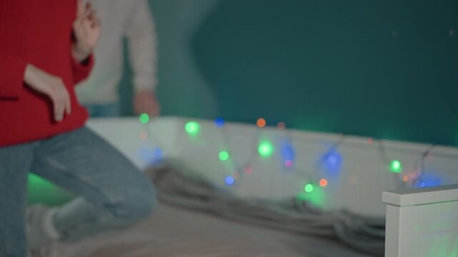 Man leans close behind woman in red sweater as they sit on bed, smiling and sharing gentle touch while colorful string lights glow along bedside panel in cozy bedroom, at eye level