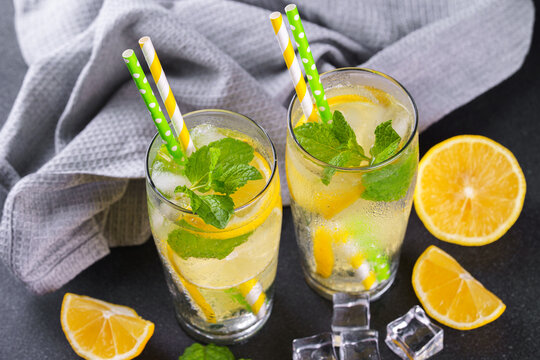 Close up of refreshing cold icy beverage, homemade lemonade with fresh lemon slices, mint leaves, ice cubes wtih soda water and condensation in glasses on black textured background