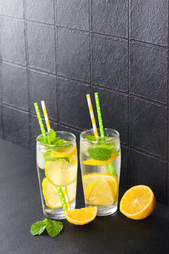 Close up of two clear glasses with refreshing homemade lemonade with fresh lemon slices, mint leaves, ice cubes and paper straws on a light kitchen counter against black tile background, copy space