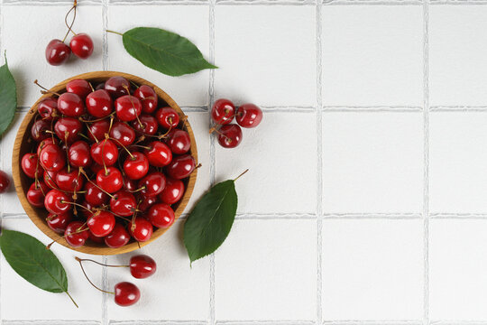 Top view of fresh ripe red sweet cherries in wooden bowl with green leaves on white tile textured background, copy space. Organic summer fruit, juicy berries, natural healthy eating, seasonal food