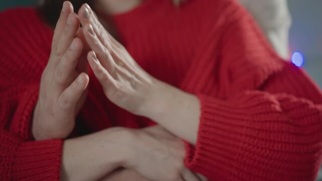 Hands remain clasped in supportive embrace while subtle shifting motion suggests ongoing comfort and reassurance, with red knit sleeves, soft indoor light and caring human connection