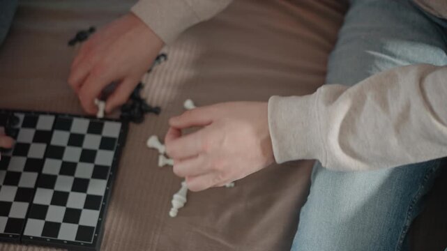 Close-up angled top-down scene of hands placing white chess pieces beside compact foldable chessboard on woven fabric blanket, fingers moving with slight blur while scattered black pieces wait nearby