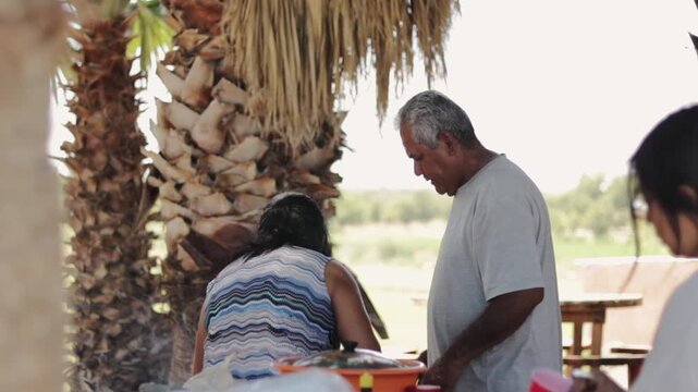 Elderly indigenous couple preparing a meal outdoors during a family gathering next to palm trees on a sunny day in a rural setting, embodying traditional lifestyle