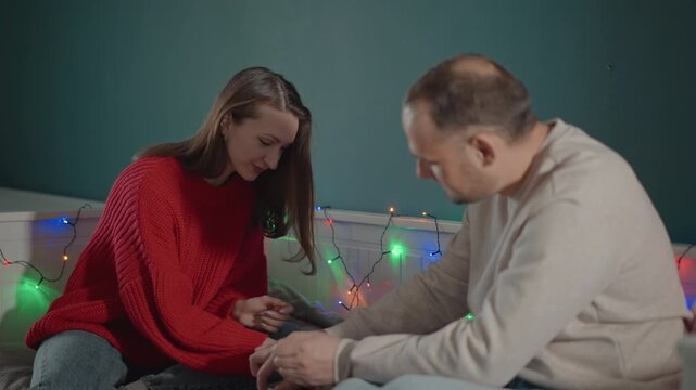 Teenage daughter and middle aged father sit on bed beside warm string lights, exchanging small wrapped present while she holds it with both hands and he reaches forward, sharing cozy celebration