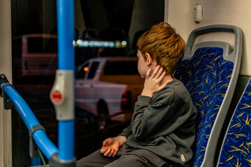 A young boy with red hair sits on a public transport vehicle (bus or tram) in the evening or at night. © Yulia