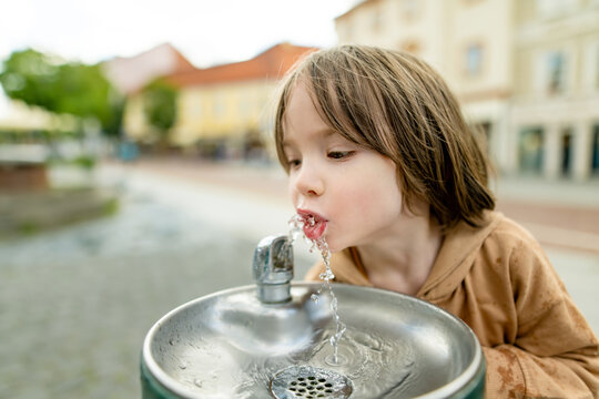 Playful young boy having fun with a public drinking fountain in the city. Laughing child splashes with fresh water outdoors during a playful urban summer moment.
