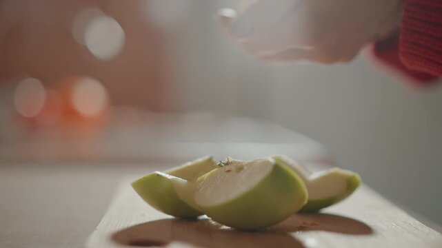 Close-up of glossy green apple wedges on wooden cutting board as blurred hand adds garnish, creating fresh domestic food preparation scene with warm light, soft bokeh, and natural texture