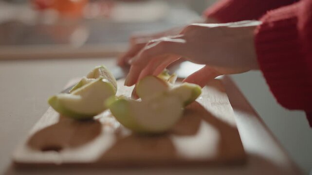 Low-angle close view of hands arranging freshly cut green apple wedges on wooden cutting board in warm kitchen, gentle serving motion visible as fingertips hover over fruit, with juice droplets
