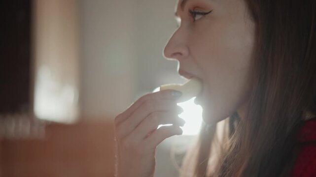 Profile view of woman wearing red sweater, eyes closed during gentle exhale beside bright kitchen window; soft backlight, rimlight, and subtle flare emphasize peaceful morning calm