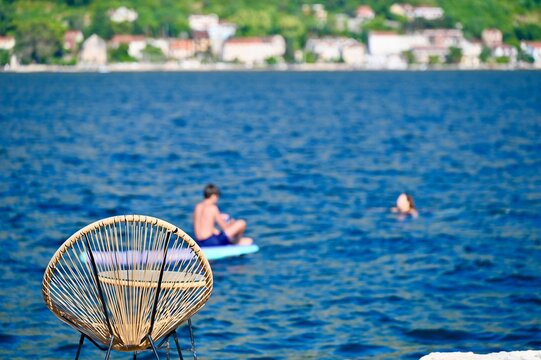 A stylish rattan chair sitting on a coastal stone shore overlooking the deep blue sea, where a person relaxes on a floating mat with a town and mountains across the water.