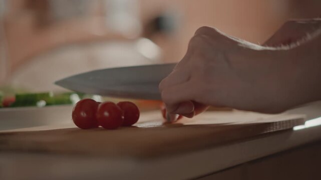 Close view of hand pressing knife through cherry tomato on wooden cutting board, steady fingers holding fruit while halves separate and juice gathers; warm kitchen light and shallow focus