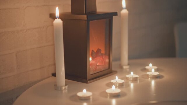 Decorative metal lantern surrounded by lit tealights and tall taper candles, with gentle flickering flames casting warm orange yellow reflections across glossy tabletop beside rustic brick wall