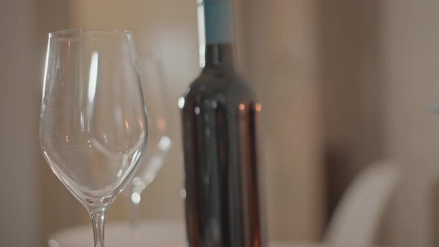 Close-up view of empty wine glass in foreground with dark bottle behind on tabletop, soft indoor light, gentle blur and droplets inside glass suggest recent movement, warm elegant dining mood