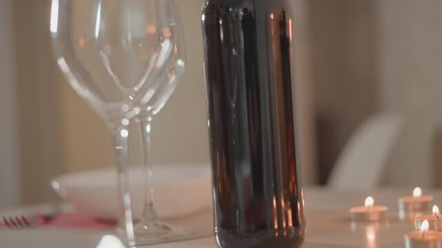 Low-angle close-up shows dark bottle flanked by two empty stemmed glasses on softly lit tabletop, with warm reflections, soft bokeh, shallow depth, and gentle blur suggesting calm movement in