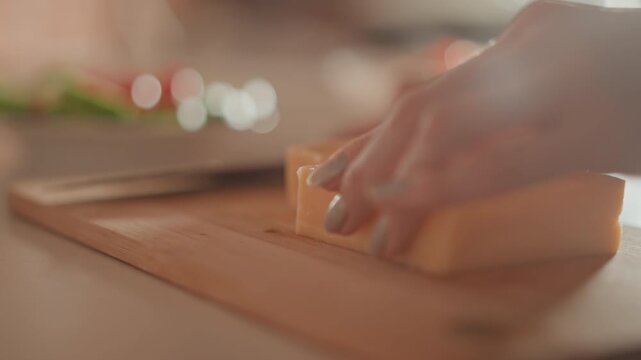 Tight kitchen scene shows hands slicing block of yellow cheese on wooden cutting board with chef knife, revealing thin even pieces, firm grip, fresh texture, and careful food prep motion