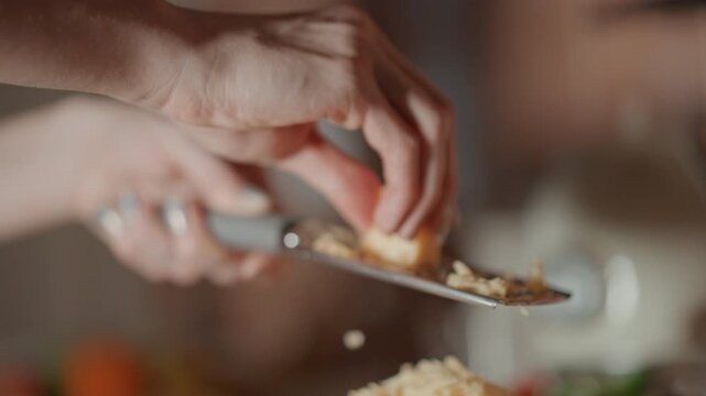 Hands pressing block of hard cheese across metal grater while steadying grater over white bowl, visible downward motion producing fine shavings that fall into bowl