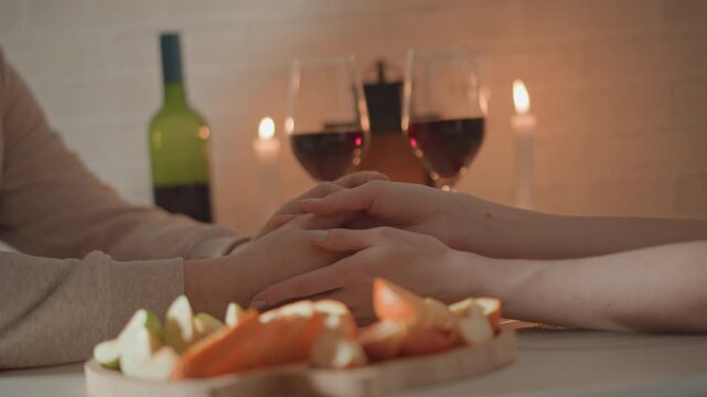 Two people hold hands across low candlelit table, fingers interlaced and palms clasped while leaning toward each other, with wine, cheese and warm ambient light creating calm intimate connection