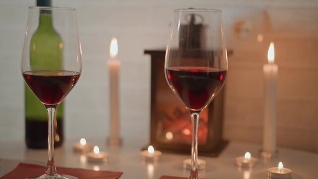 Close-up of single red wine glass on tabletop with glowing candle reflections, subtle ripple on surface, blurred wine bottle, taper candles, fireplace light and refined dinner setting