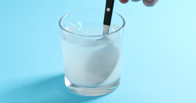 Woman pouring baking soda into glass and stirring it on light blue background, closeup