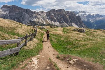 Fototapeta premium Young woman hiking away with her dog along a narrow dirt trail on a grassy mountain ridge next to a rustic wooden fence, spectacular alpine rock formations concept
