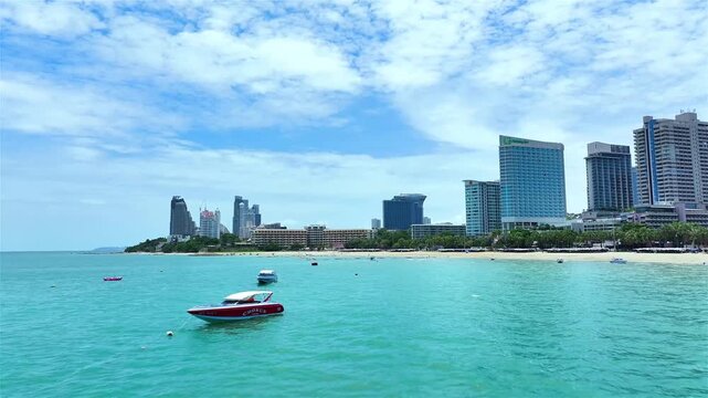 An aerial view of the Pattaya coastline with modern high-rise hotels and condos along the turquoise sea and a passenger boat visible.