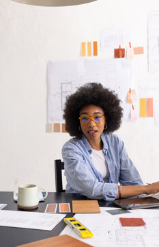 Adult African American woman in blue shirt, yellow glasses reviewing plans at desk with swatches