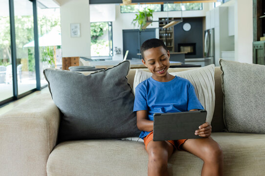 African boy sitting on sofa in living room holding black tablet wearing blue tee, orange shorts